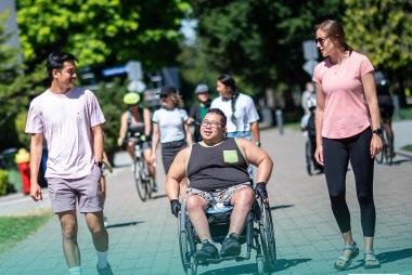 Students walking and chatting, one in a wheelchair, on a sunny campus pathway.